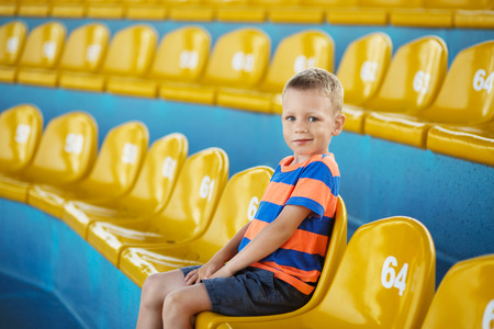 Little boy sitting in an empty stadium among yellow plastic numbered seats and applauds. Child take own seat in the stadium or dolphinarium and waiting patiently when the show or game starts.の写真素材