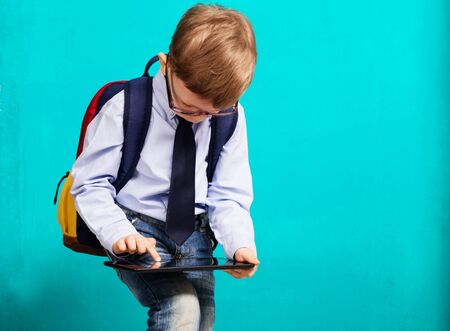 Cheerful little boy with big backpack holding digital tablet against blue background. Child playing games on a touch pad. School concept. Back to Schoolの写真素材