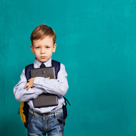 Cheerful little boy with big backpack holding digital tablet against blue background. Looking at camera. School concept. Back to Schoolの写真素材