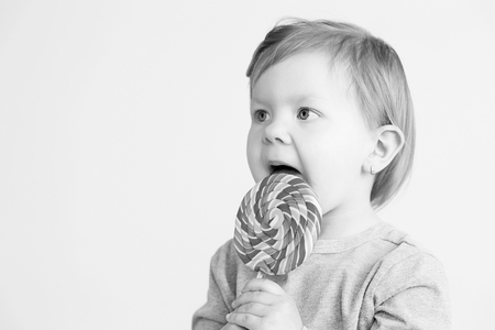 Small girl eating lollipop. Happy children with a big delicious candy. Portrait of little baby girl against a white background. Black and white photographyの写真素材