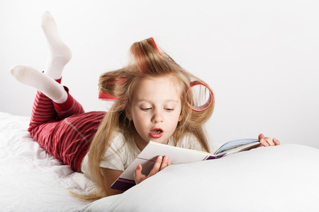 Beautiful fashion little girl with curlers on her head reading a book Lying on the bed at home. Day off, holiday, day of rest, free, off-time.の写真素材