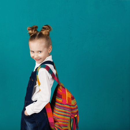 Beautiful smiling little girl with big Knitted backpack standing against blue wall. Looking at...