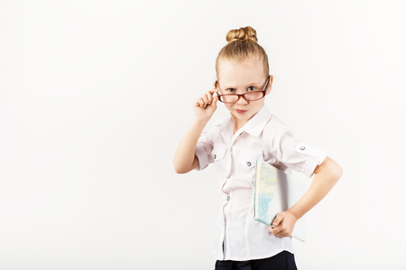 Funny little girl wearing eyeglasses imitates a strict teacher against white background.  Little student holding books.  Looking at camera.  School concept.  Back to Schoolの写真素材