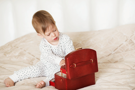 Little girl admires the accessories in the box. Baby girl playing with her mother's jewelry sitting on a bed in pajamas barefootの写真素材