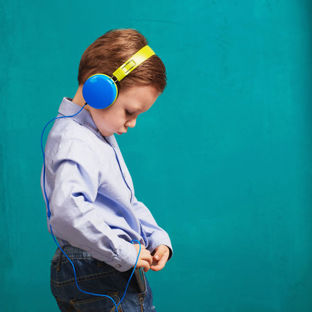 Handsome little stylish boy in bright headphones standing and listening to music on headphones via...