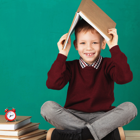 Cheerful smiling little school boy with big heavy books on his head sitting on a desk against turquoise wall. Looking at camera. School concept. Back to Schoolの写真素材