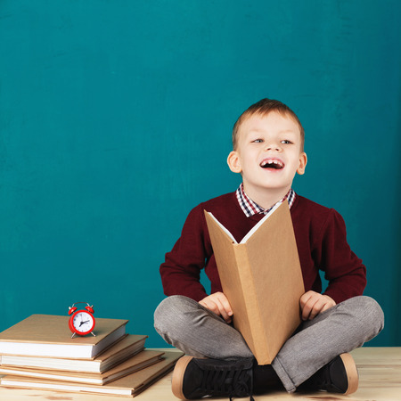 Cheerful smiling little school boy sitting on a desk against turquoise wall and reading book. Looking at camera. School concept. Back to Schoolの写真素材