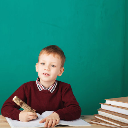smiling little schoolboy writing notes in class against green wall. A child draws in a notebook with...