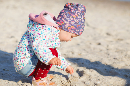 Cute little girl playing on the sandy beach. Happy child wearing warm floral print jacket, pom pom hat and scarf playing outdoors on fall, winter or spring day. Family enjoying winter walk in nature.の写真素材