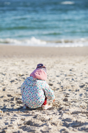 Cute little girl playing on the sandy beach. Happy child wearing warm floral print jacket, pom pom hat and scarf playing outdoors on fall, winter or spring day. Family enjoying winter walk in nature.の写真素材
