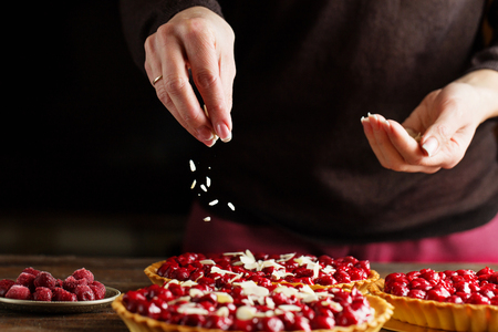Beautiful freshly made cherry tart decorated with almond chips. Food photography. Process of making tart. Close up hands of the chef sprinkling cake with almond chips at pastry shop kitchen.の写真素材