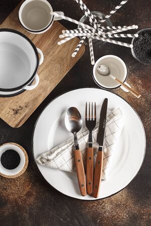 Crockery and cutlery on a dark background with copy space. White metal plates, bowls, cups and cutlery with wooden handles. Set of dishesの写真素材