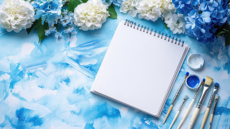 An open sketchbook on a soft blue watercolor backdrop, with brushes, paints, and white and blue flowers, suggesting the creative flow of artistic endeavor.の素材
