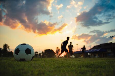 Silhouette action sport outdoors of a group of kids having fun playing soccer football on green grass field for exercise in community rural area under the twilight sunset sky.の写真素材