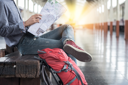 Side portrait of a young man traveler sitting with map choose where to travel and bag waiting for train at train station.の写真素材