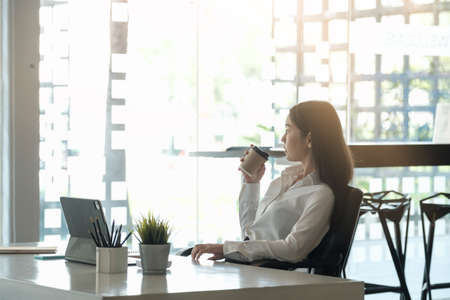 Portrait of beautiful woman or accountant sitting at desk in modern office with interior drinking hot beverage holding cup with coffee in hands looking outside enjoying freetime.の写真素材