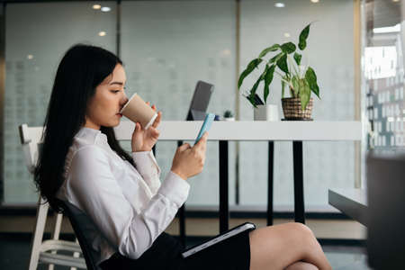 Businesswomen using smartphone read and text message while holding cup coffee at her office.の写真素材