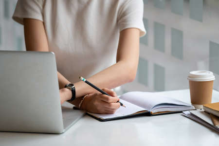 Cropped photo hand of woman writing making list taking notes in notepad working or learning online with laptop at homeの写真素材