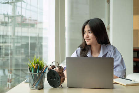 Portrait young asian girl looking at window outside while using laptop computer for online learningの写真素材