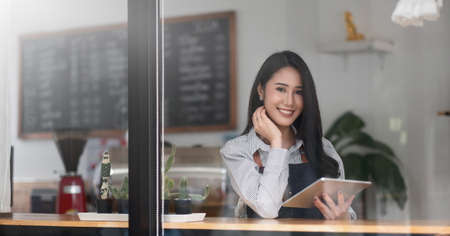 Cheerful young asian woman owner holding digital tablet while standing in her cafe, young...