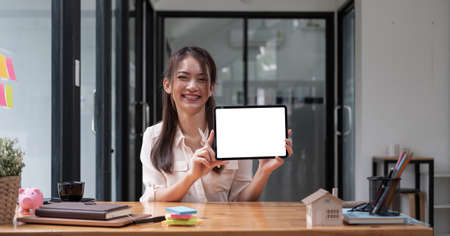 Portrait of Asian woman showing or presenting tablet computer with blank white screen, real estate working at officeの写真素材