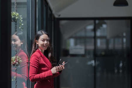 Image of young asian woman holding mobile phone while working in office, she looking at cameraの写真素材