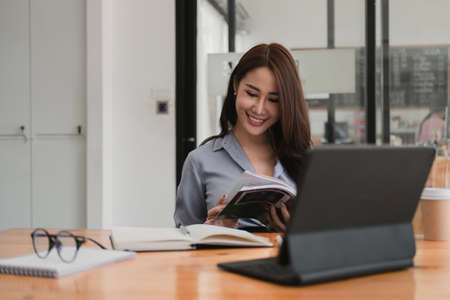 Smiling asian woman reading a book while studying or webinar online via tablet, video conference conceptの写真素材