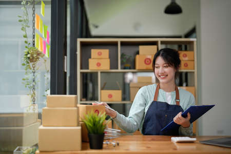 A portrait of Asian woman, e-commerce employee sitting in the office full of packages on the table using a calculator, for SME business, e-commerce, technology and delivery businessの写真素材