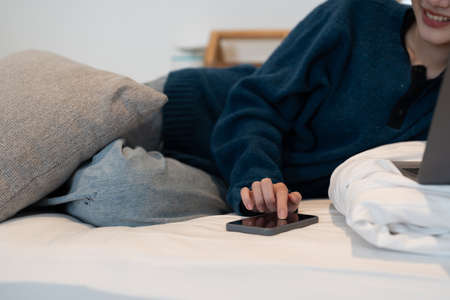 Happy young asian woman using mobile phone while sitting a couch at home with laptop computer.の写真素材