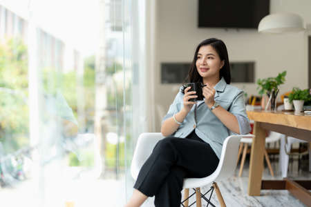 Beautiful asian businesswoman in elegance suit sitting at office desk and holding a coffee cup. Female entrepreneur, business owner.の写真素材