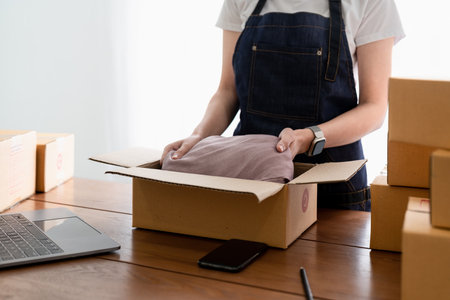 Young woman working online shopping and packing clothes with cardboard box at home - Business online...