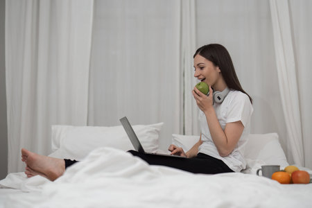 Young woman relaxing and eat fruit using laptop on checking social apps and working the bedroomの写真素材