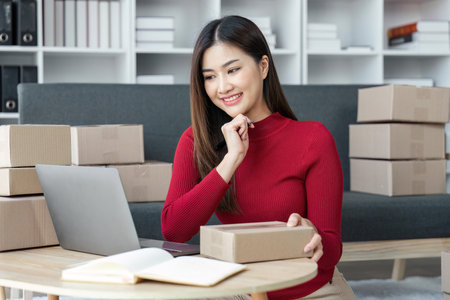Female seller write the customers address on the parcel box to prepare for delivery and using laptop...