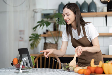 Image of young pretty lady in kitchen and cooking the salad. Looking at tablet computerの写真素材