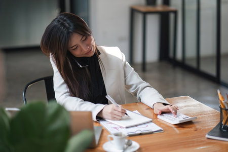 businesswoman or accountant taking note and working on calculator and laptop computer to calculate business data during make note at notepad, accountancy document at office.の写真素材