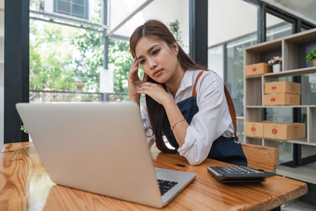 Small online business owner Asian woman wearing apron stressed headache taking notes with parcel box at homeの写真素材