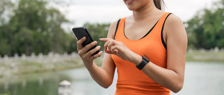 Portrait of smiling asian woman looking at her smartphone and reading on application, standing in park wearing sport uniformの写真素材