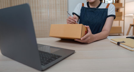 Woman who runs an ecommerce business is writing a list of customers on box before shipping to them,...