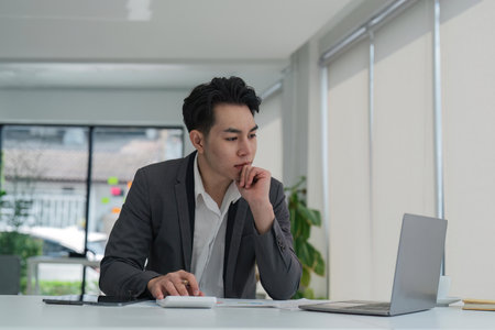 Focused young adult businessman asian sitting at desk making sure his company is earning enough money financial planning conceptの写真素材