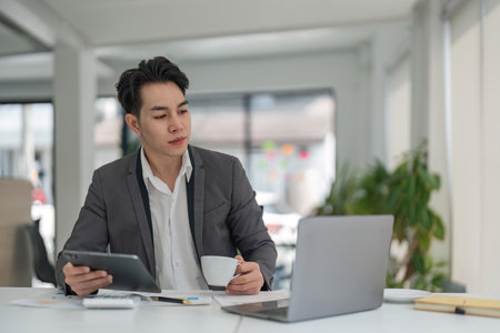 Focused young adult businessman asian sitting at desk making sure his company is earning enough...