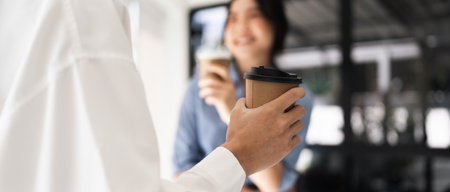 Couple of happy colleagues talking while standing at the office window with cups of coffee