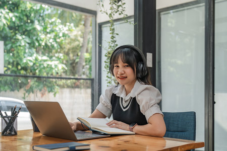 Smiling young woman in wireless headphones sitting at desk table working on laptop and writing in paper notebookの写真素材