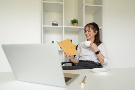 Beautiful young Asian businesswoman drinking a coffee and holding a book working on laptop at office