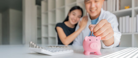 Family savings. Happy couple asian putting coin in piggybank while sitting together on desk at home, loving spouse planning budget and saving money for futureの写真素材