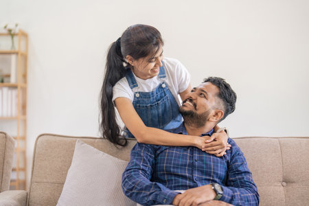 Happy lovely young Indian couple together at home, young wife hugging from behind her husband, sitting and rest on sofa in home, portrait of romantic multiracial couple in loveの写真素材