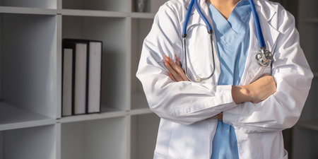 Friendly asian woman doctor in workwear with stethoscope on neck posing with folded arms in clinic interiorの写真素材