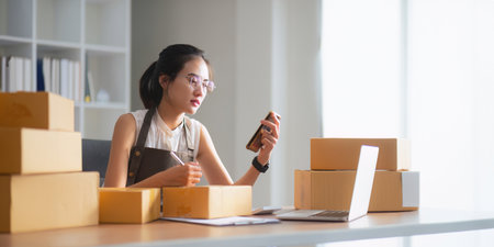 Startup small business entrepreneur of freelance Asian woman using smartphone and box to receive and review order online to prepare to pack sell to customers, online business ideasの写真素材