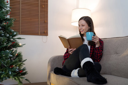 Young cute woman holding cup coffee or tea and read interest book. Concept of Christmas atmosphere at homeの写真素材