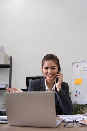 Cheerful young asian business woman talking on phone working in office. Happy positive Asian businesswoman company manager wearing suit making call on cellphone sitting at workplaceの写真素材