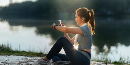 Portrait of smiling Asian woman looking at her smartphone and watch, standing in park wearing sport uniformの写真素材